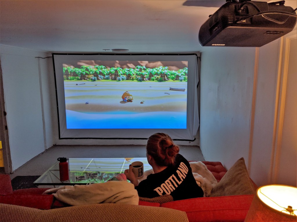 Basement with large screen on far war with a video game image projected on it. Woman sitting on couch playing the game.