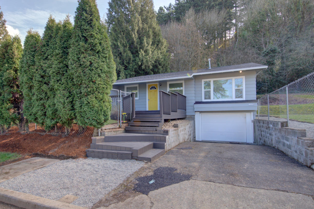 Exterior gray house with a yellow door, driveway and several treess