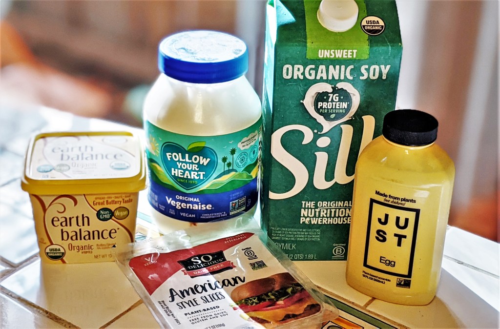 A group of vegan products: Earth Balance, Vegenaise, Silk Soy Milk, Just Egg and fake cheese slices in a plastic container.