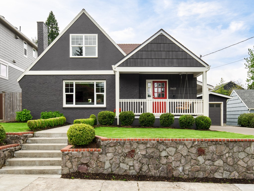 Exterior of dark gray home with red door and covered front porch. Green grass.
