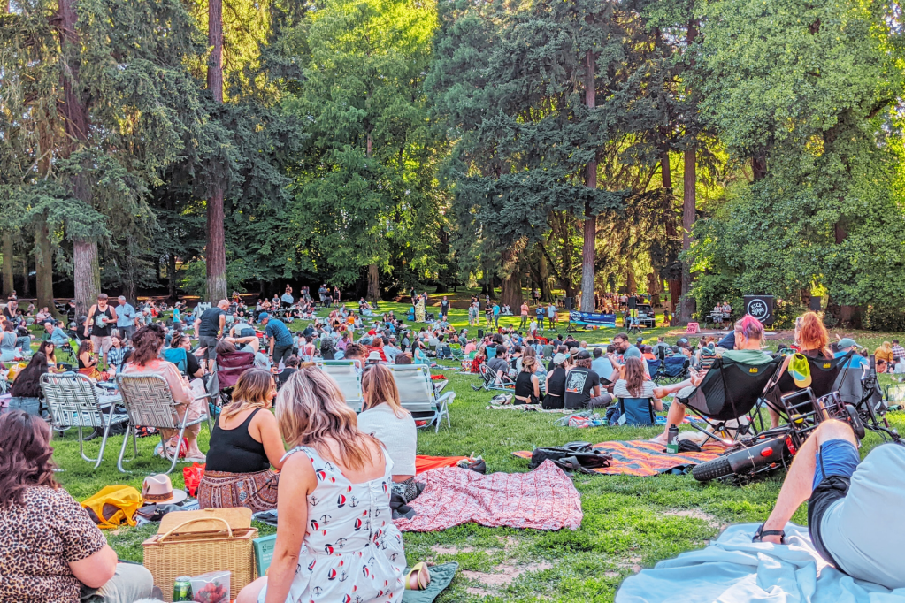 A crowd of people seated on blankets at Laurelhurst Park surrounded by trees
