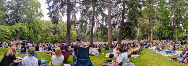 Crowd of people sitting on the grass under the trees at Laurelhurst Park watching a comedian perform in the distance